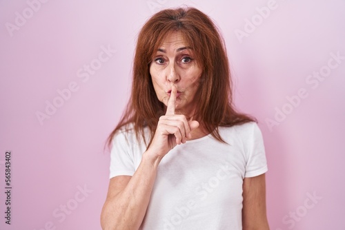 Middle age woman standing over pink background asking to be quiet with finger on lips. silence and secret concept.