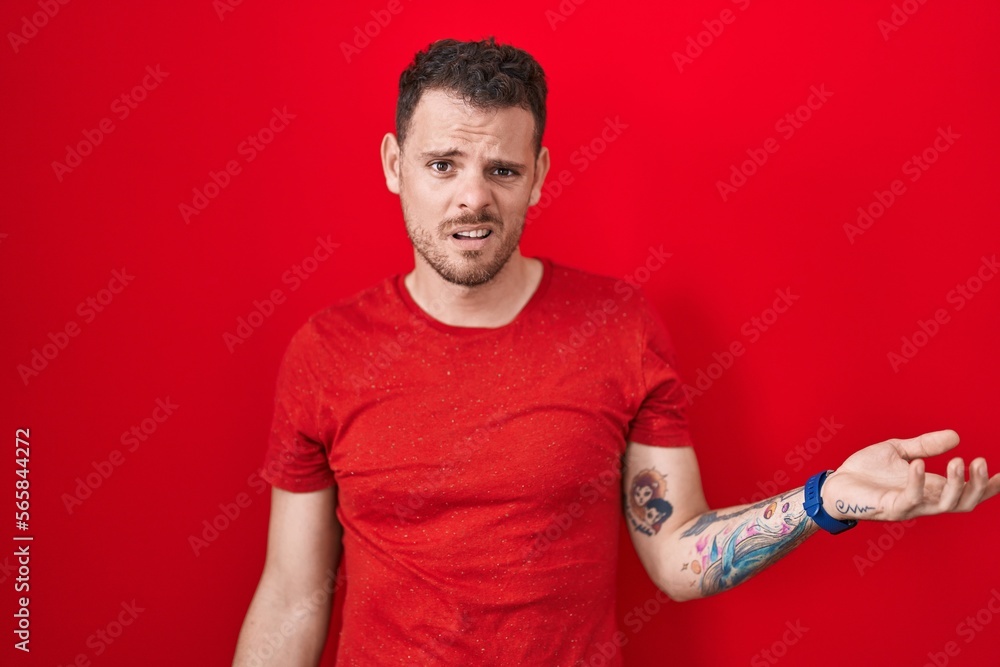 Young hispanic man standing over red background in shock face, looking skeptical and sarcastic, surprised with open mouth Young hispanic man standing over red background in shock face, looking skeptical and sarcastic, surprised with open mouth