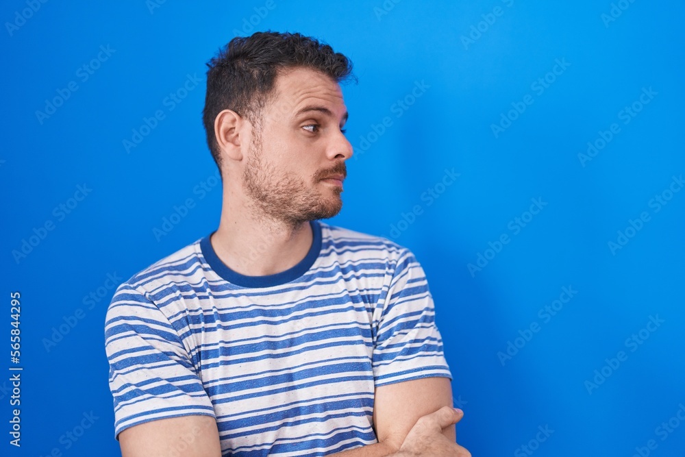 Young hispanic man standing over blue background looking to the side with arms crossed convinced and confident Young hispanic man standing over blue background looking to the side with arms crossed convinced and confident