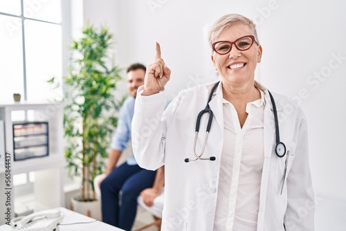 Female doctor wearing uniform and stethoscope at medical clinic smiling with an idea or question pointing finger with happy face, number one