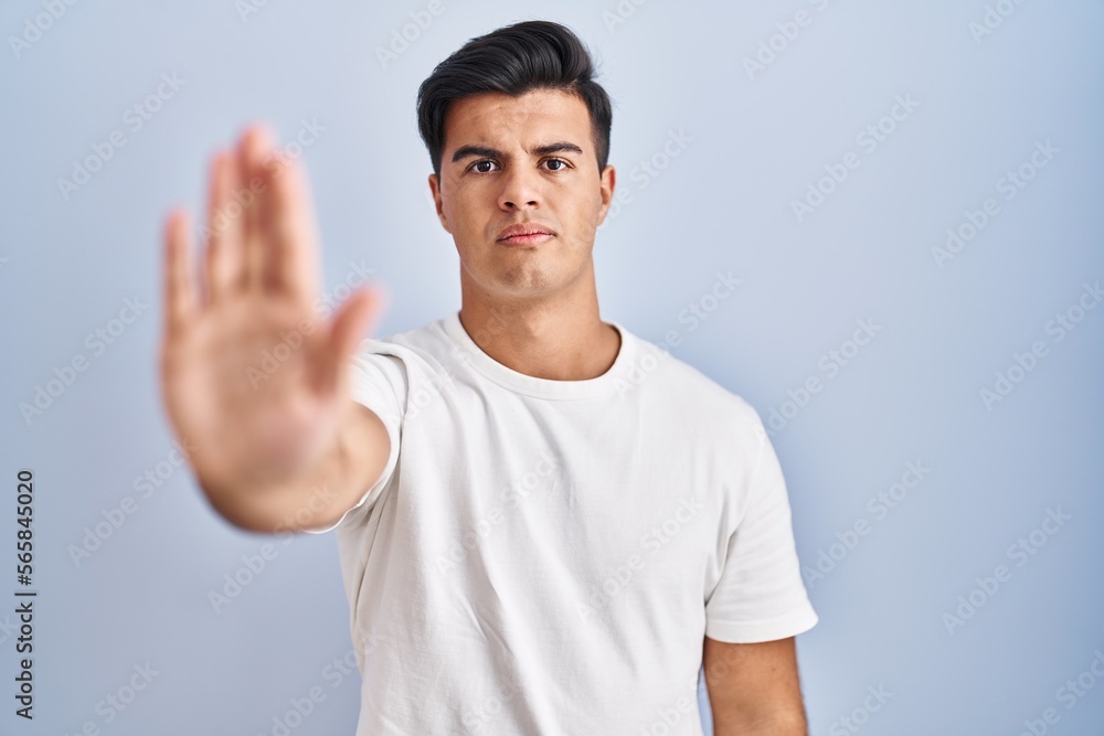 Hispanic man standing over blue background doing stop sing with palm of the hand. warning expression with negative and serious gesture on the face. Hispanic man standing over blue background doing stop sing with palm of the hand. warning expression with negative and serious gesture on the face.