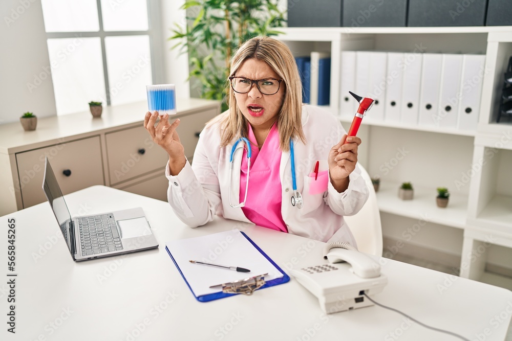 Young hispanic doctor woman holding ear otoscope and cotton buds angry and mad screaming frustrated and furious, shouting with anger. rage and aggressive concept. Young hispanic doctor woman holding ear otoscope and cotton buds angry and mad screaming frustrated and furious, shouting with anger. rage and aggressive concept.