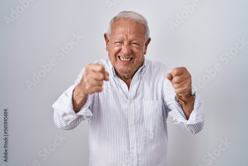 Senior man with grey hair standing over isolated background angry and mad raising fists frustrated and furious while shouting with anger. rage and aggressive concept.