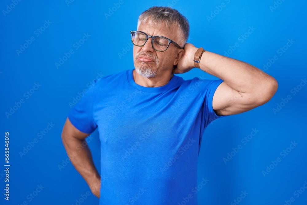 Hispanic man with grey hair standing over blue background suffering of neck ache injury, touching neck with hand, muscular pain Hispanic man with grey hair standing over blue background suffering of neck ache injury, touching neck with hand, muscular pain