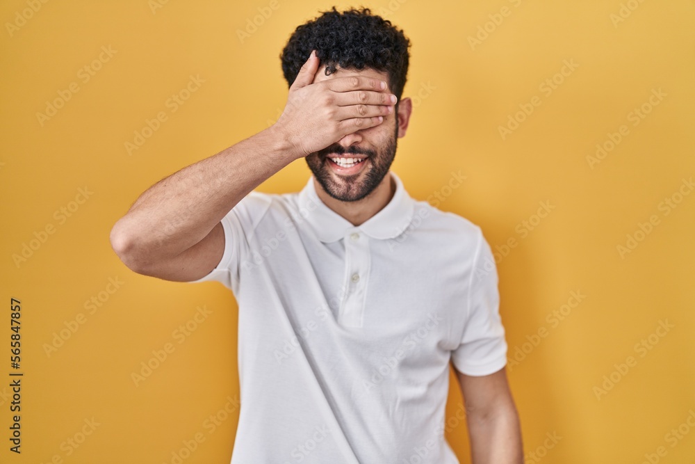 Arab man standing over yellow background smiling and laughing with hand on face covering eyes for surprise. blind concept. Arab man standing over yellow background smiling and laughing with hand on face covering eyes for surprise. blind concept.