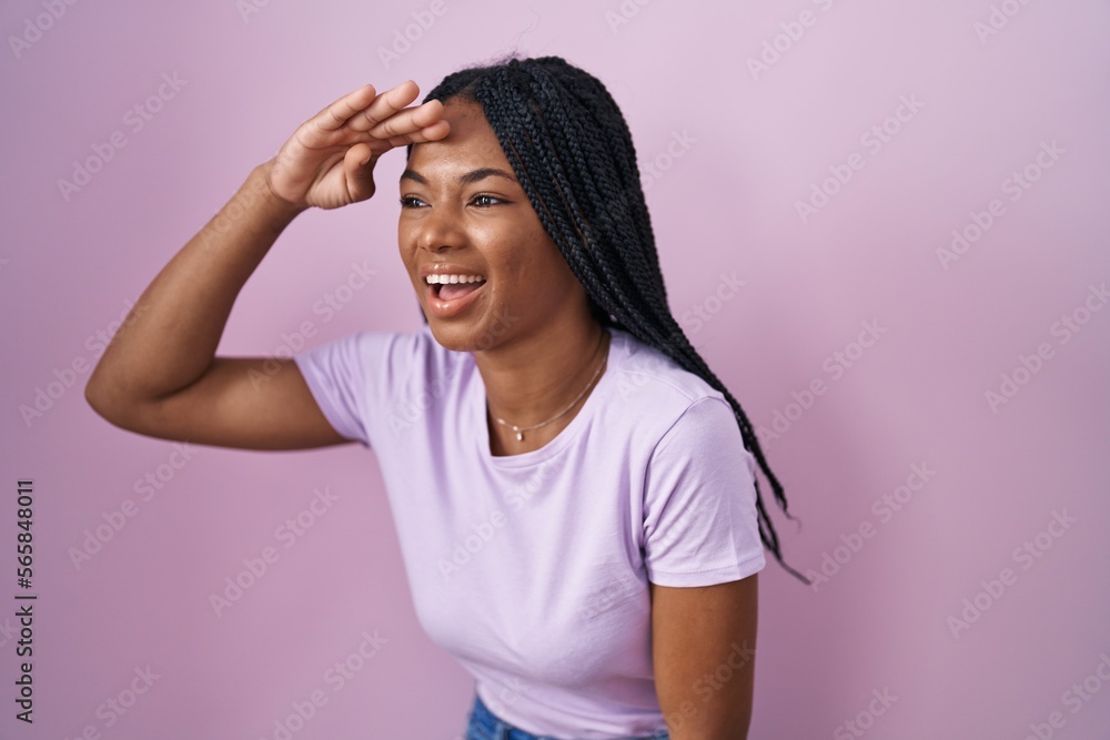 African american woman with braids standing over pink background very happy and smiling looking far away with hand over head. searching concept. African american woman with braids standing over pink background very happy and smiling looking far away with hand over head. searching concept.