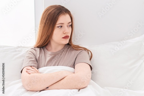 Young redhead woman sitting on bed with unhappy expression and arms crossed gesture at bedroom