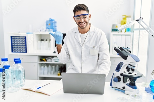 Young hispanic man working at scientist laboratory with laptop pointing thumb up to the side smiling happy with open mouth
