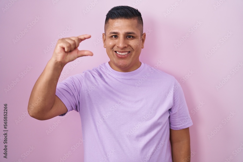 Young hispanic man standing over pink background smiling and confident gesturing with hand doing small size sign with fingers looking and the camera. measure concept. Young hispanic man standing over pink background smiling and confident gesturing with hand doing small size sign with fingers looking and the camera. measure concept.