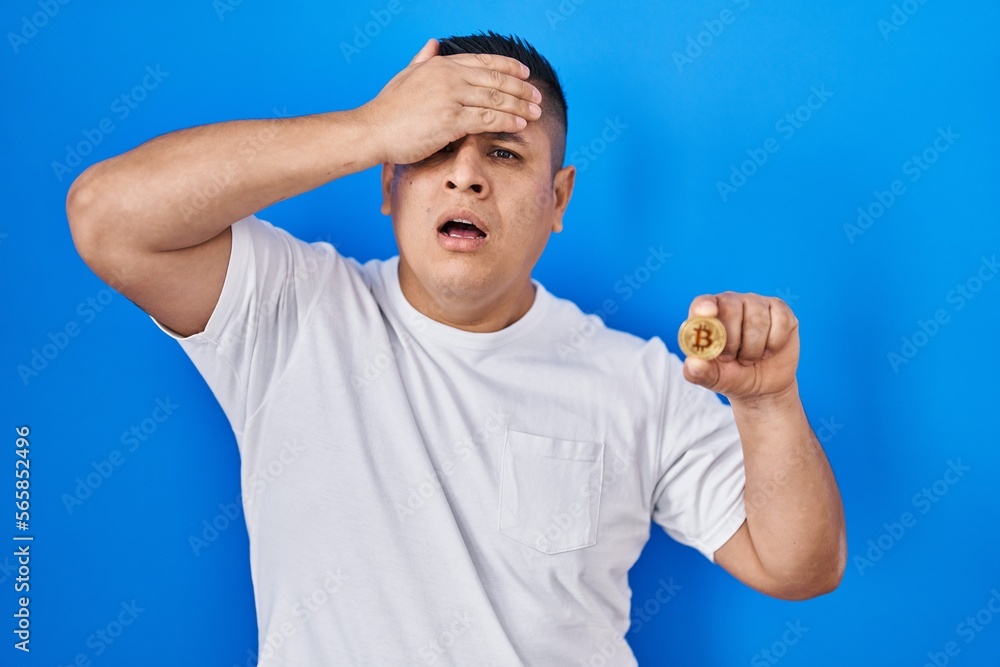 Hispanic young man holding virtual currency bitcoin stressed and frustrated with hand on head, surprised and angry face Hispanic young man holding virtual currency bitcoin stressed and frustrated with hand on head, surprised and angry face