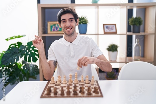 Young hispanic man playing chess sitting on the table smiling happy pointing with hand and finger to the side