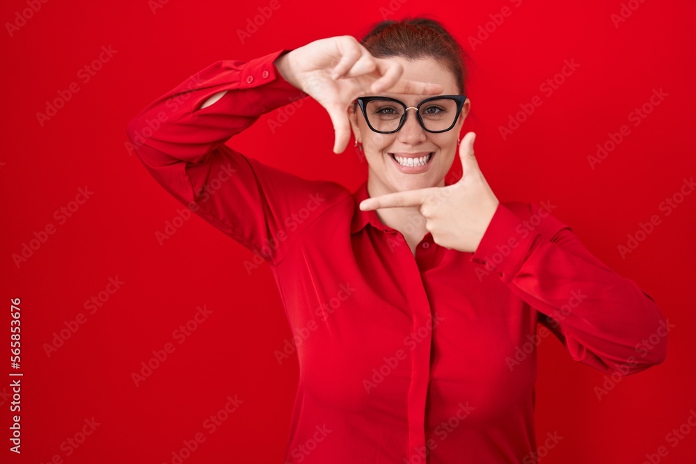 Obraz premium Young hispanic woman with red hair standing over red background smiling making frame with hands and fingers with happy face. creativity and photography concept.