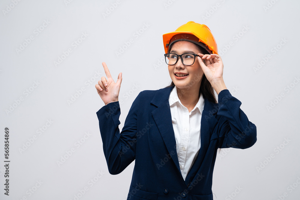 Portrait of Asian female engineer with hard hat pointing at copy space isolated on grey background.