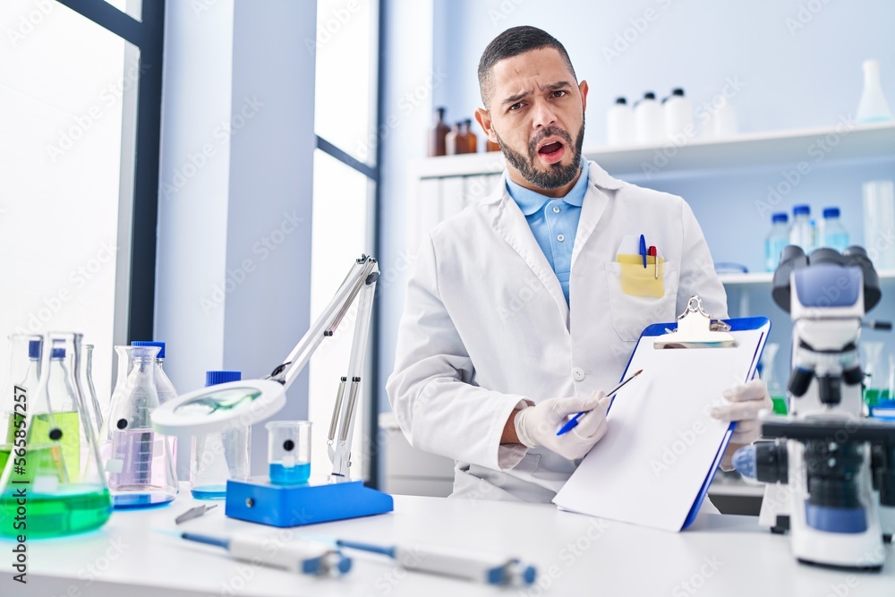 Hispanic man working at scientist laboratory holding blank clipboard in shock face, looking skeptical and sarcastic, surprised with open mouth Hispanic man working at scientist laboratory holding blank clipboard in shock face, looking skeptical and sarcastic, surprised with open mouth