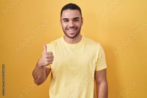 Young hispanic man standing over yellow background doing happy thumbs up gesture with hand. approving expression looking at the camera showing success.