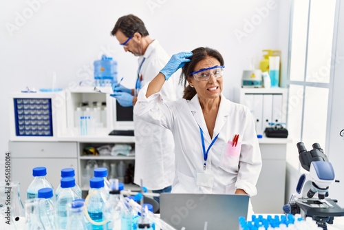 Middle age woman working at scientist laboratory stressed and frustrated with hand on head  surprised and angry face