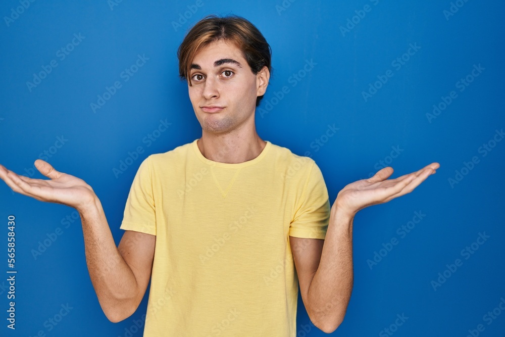 Young man standing over blue background clueless and confused expression with arms and hands raised. doubt concept. Young man standing over blue background clueless and confused expression with arms and hands raised. doubt concept.