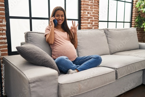 Young pregnant woman having conversation speaking on the smartphone doing ok sign with fingers, smiling friendly gesturing excellent symbol