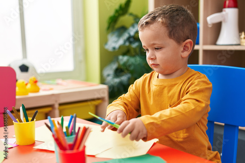 Adorable caucasian boy student cutting paper sitting on table at kindergarten