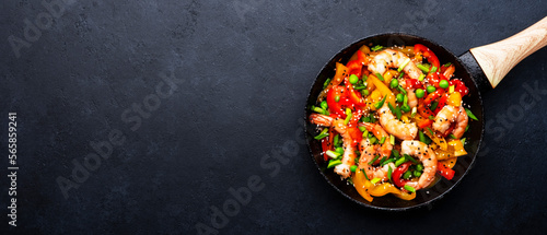 Fototapeta Naklejka Na Ścianę i Meble -  Stir fry with shrimps, red and yellow paprika, green pea, chives and sesame seeds in frying pan. Asian cuisine dish. Black stone kitchen table background, top view banner