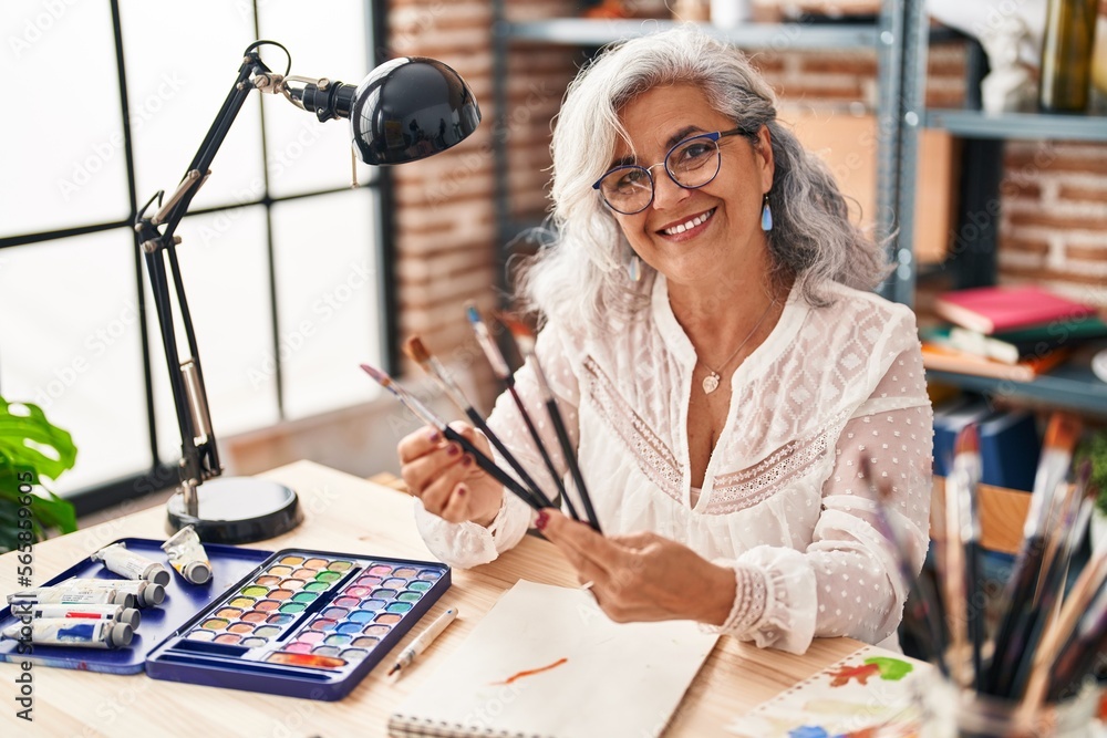 Fototapeta premium Middle age woman artist smiling confident holding paintbrushes at art studio