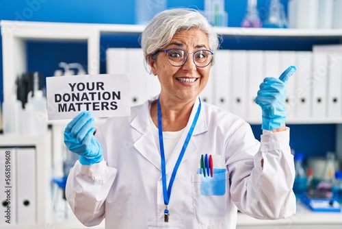 Middle age woman with grey hair working at scientist laboratory holding your donation matters banner smiling happy pointing with hand and finger to the side