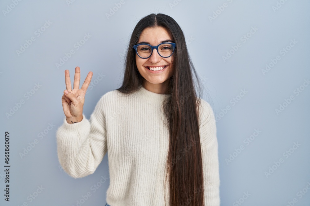 Young hispanic woman wearing casual sweater over blue background showing and pointing up with fingers number three while smiling confident and happy. Young hispanic woman wearing casual sweater over blue background showing and pointing up with fingers number three while smiling confident and happy.