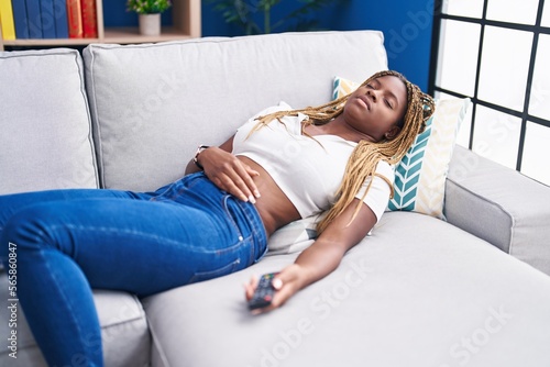 African american woman holding tv remote control lying on sofa sleeping at home