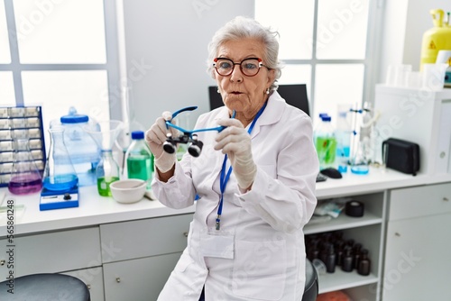 Senior woman with grey hair working at scientist laboratory using magnifying glasses making fish face with mouth and squinting eyes, crazy and comical.