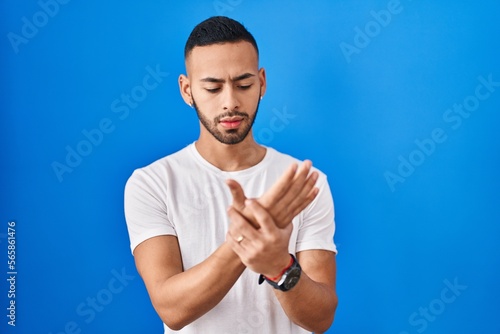 Young hispanic man standing over blue background suffering pain on hands and fingers, arthritis inflammation