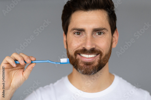 Excited handsome man holding toothbrush, standing over grey studio baground and smiling at camera