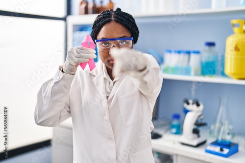 African american woman working at scientist laboratory holding pink ribbon pointing with finger to the camera and to you  confident gesture looking serious