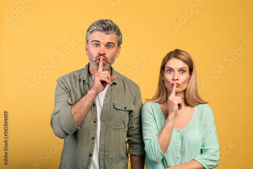 Keep silence concept. Middle aged spouses gesturing hush sign and looking at camera, standing over yellow background