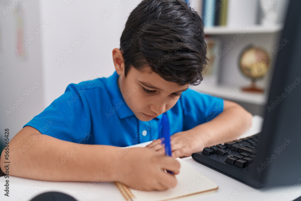 Adorable hispanic boy student using computer writing on notebook at classroom Adorable hispanic boy student using computer writing on notebook at classroom