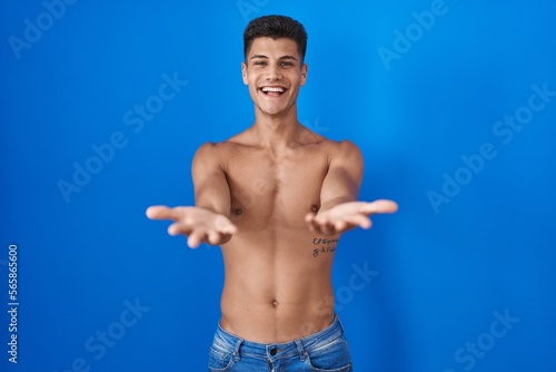 Young hispanic man standing shirtless over blue background smiling cheerful offering hands giving assistance and acceptance.