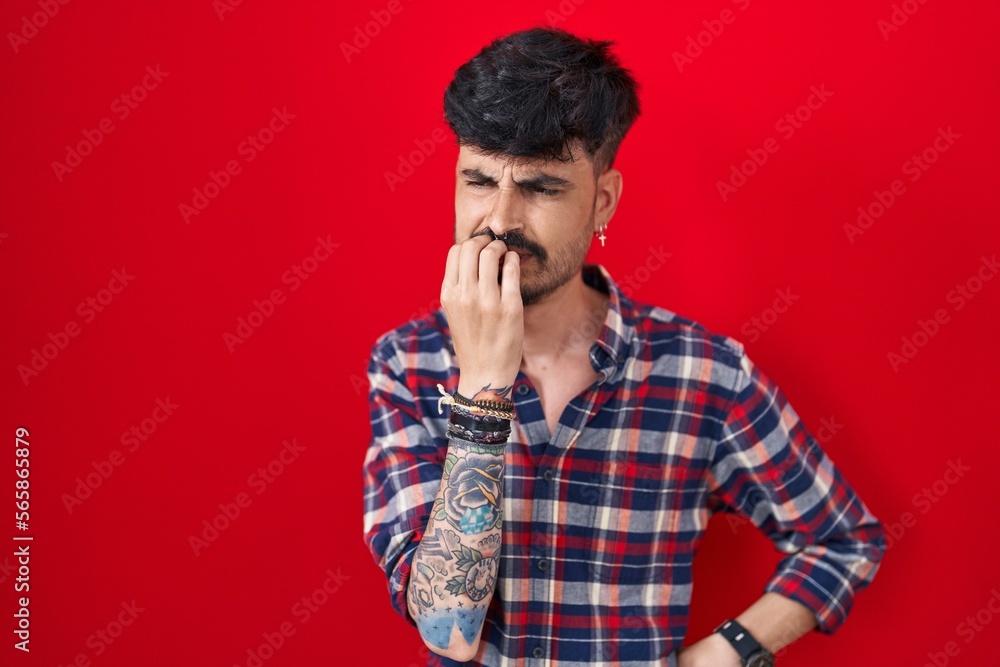 Young hispanic man with beard standing over red background looking stressed and nervous with hands on mouth biting nails. anxiety problem.