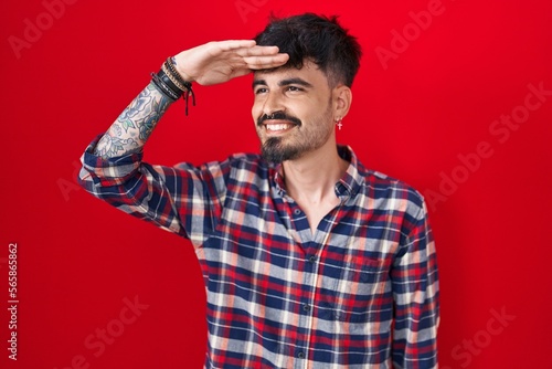 Young hispanic man with beard standing over red background very happy and smiling looking far away with hand over head. searching concept. © Krakenimages.com