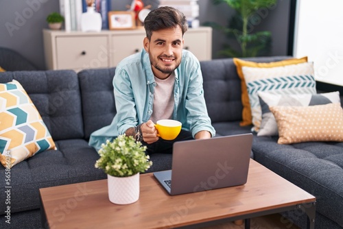 Young hispanic man using laptop drinking coffee at home