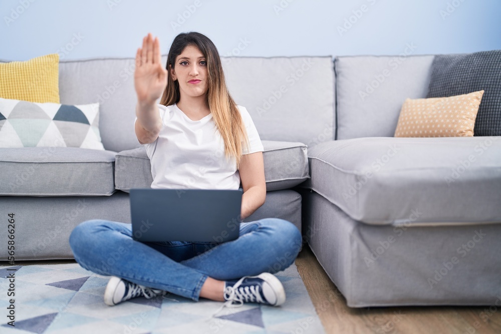 Young brunette woman sitting on the floor at home using laptop with open hand doing stop sign with serious and confident expression, defense gesture Young brunette woman sitting on the floor at home using laptop with open hand doing stop sign with serious and confident expression, defense gesture
