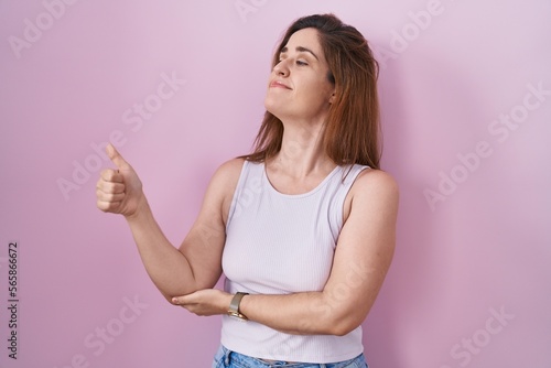 Brunette woman standing over pink background looking proud, smiling doing thumbs up gesture to the side © Krakenimages.com