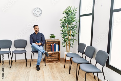 Young hispanic man using smartphone sitting on chair at waiting room