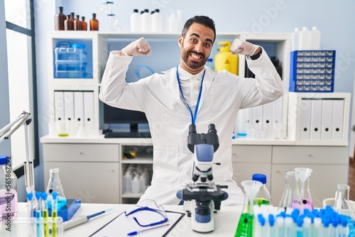 Young hispanic man with beard working at scientist laboratory showing arms muscles smiling proud. fitness concept.
