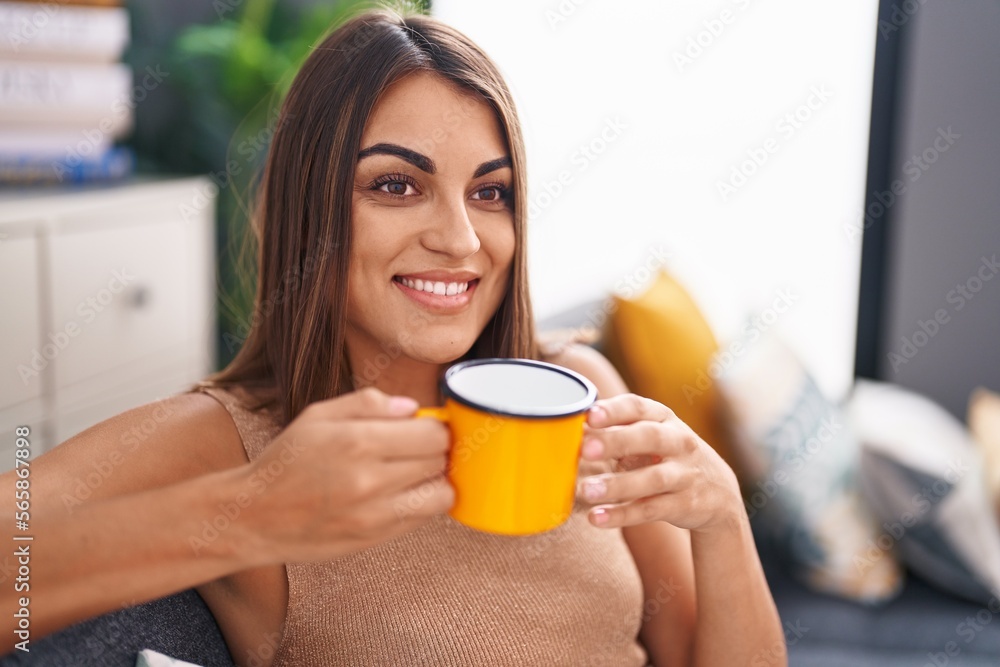 Young beautiful hispanic woman drinking coffee sitting on sofa at home