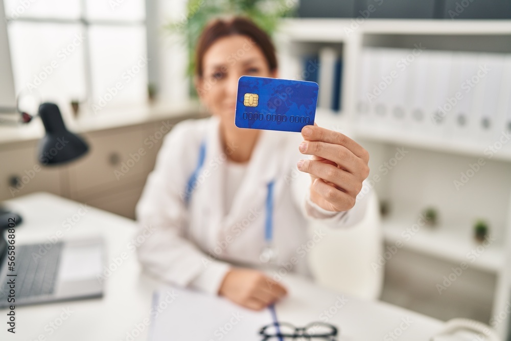 Young hispanic woman wearing doctor uniform holding credit card at clinic