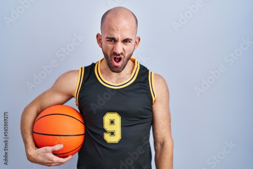 Young bald man with beard wearing basketball uniform holding ball in shock face, looking skeptical and sarcastic, surprised with open mouth