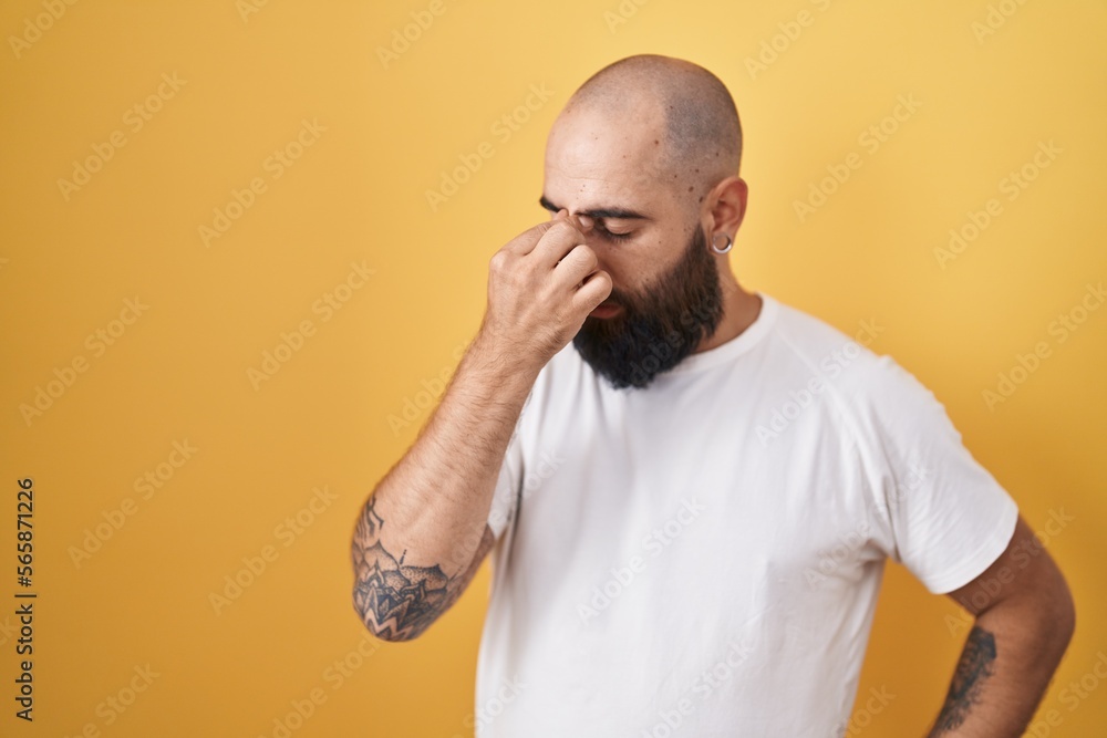 Young hispanic man with beard and tattoos standing over yellow background tired rubbing nose and eyes feeling fatigue and headache. stress and frustration concept. Young hispanic man with beard and tattoos standing over yellow background tired rubbing nose and eyes feeling fatigue and headache. stress and frustration concept.