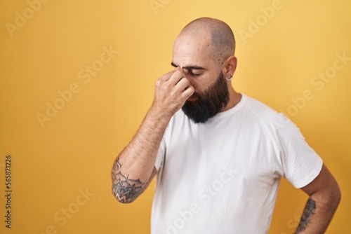 Young hispanic man with beard and tattoos standing over yellow background tired rubbing nose and eyes feeling fatigue and headache. stress and frustration concept. © Krakenimages.com