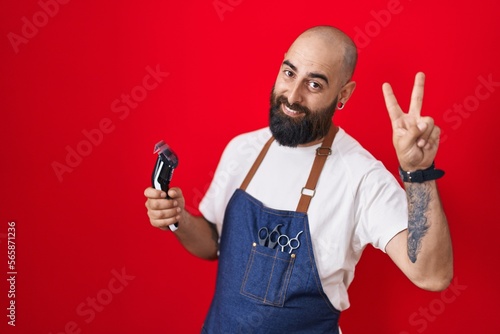Young hispanic man with beard and tattoos wearing barber apron holding razor smiling looking to the camera showing fingers doing victory sign. number two.
