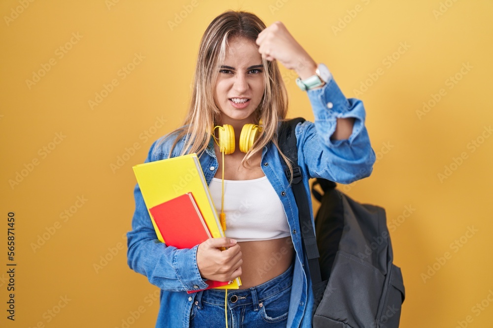 Young blonde woman wearing student backpack and holding books angry and mad raising fist frustrated and furious while shouting with anger. rage and aggressive concept. Young blonde woman wearing student backpack and holding books angry and mad raising fist frustrated and furious while shouting with anger. rage and aggressive concept.