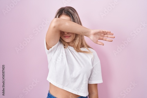 Young blonde woman standing over pink background covering eyes with arm, looking serious and sad. sightless, hiding and rejection concept